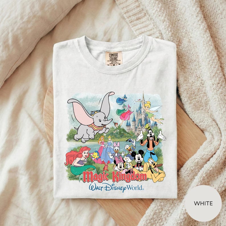 A white t-shirt with a colorful graphic design depicting various Disney characters from the Magic Kingdom, including Dumbo, Minnie Mouse, Mickey Mouse, Donald Duck, and other iconic figures.