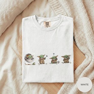 A white t-shirt with a graphic design depicting three baby alien characters in various poses, including one holding a question mark.