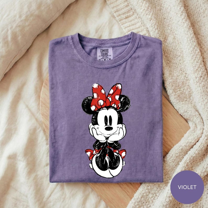 A purple t-shirt with a Minnie Mouse graphic printed on it, placed on a wooden surface.