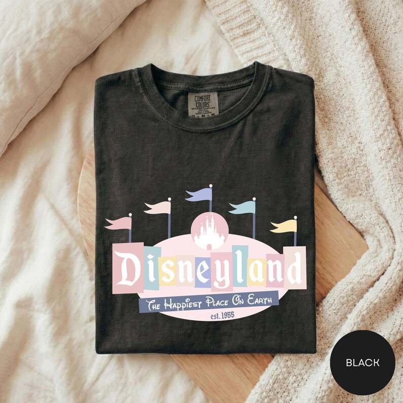 A black t-shirt with a Disneyland logo and text, laid out on a bed with a white blanket.