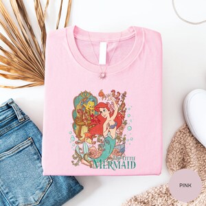 A pink t-shirt with a cartoon image of the mermaid character from the Disney film "The Little Mermaid" is displayed on a white surface, along with some jeans and a beige sweater.