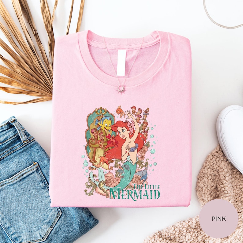A pink t-shirt with a cartoon image of the mermaid character from the Disney film "The Little Mermaid" is displayed on a white surface, along with some jeans and a beige sweater.