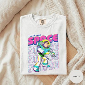 A white t-shirt with a colorful graphic design featuring a character from the animated film "Toy Story" and the text "I need some SPACE".