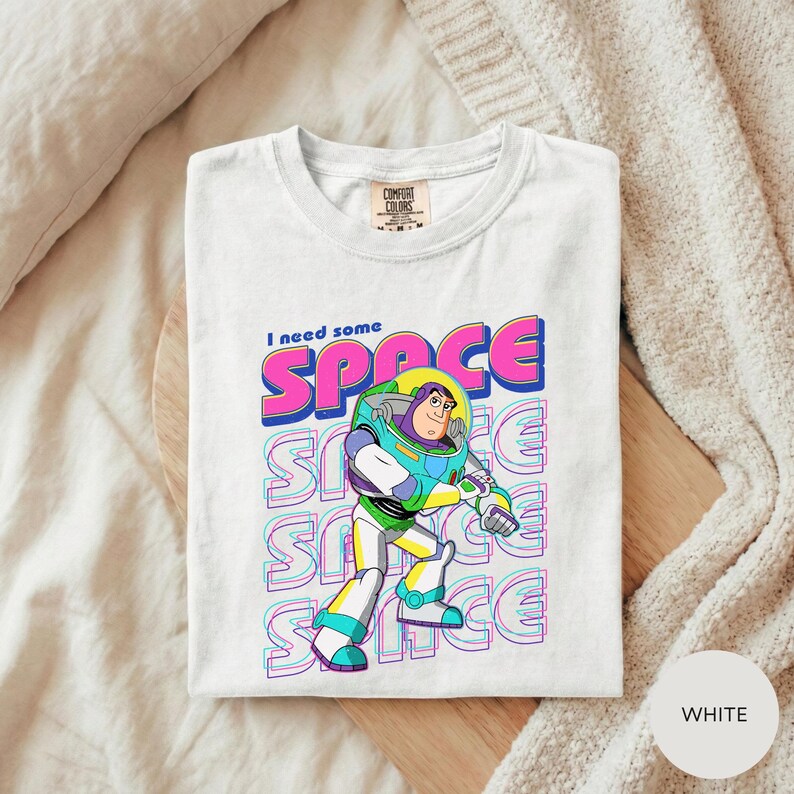 A white t-shirt with a colorful graphic design featuring a character from the animated film "Toy Story" and the text "I need some SPACE".