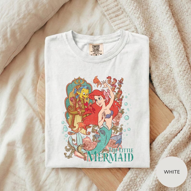 A white t-shirt with a colorful graphic design featuring characters from the Disney animated film "The Little Mermaid".