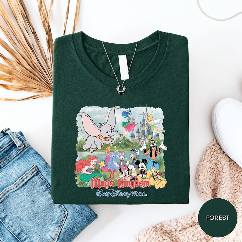 a green t-shirt with a Disney-themed graphic, including characters from the Disney film "Magic Kingdom" and a castle. The t-shirt is laid out on a white surface, along with a pair of jeans and a beige sweater.