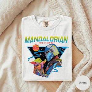 A white t-shirt with a graphic design featuring a character from the Star Wars series, specifically Mandalorian, and the text "This is the way" printed on it.