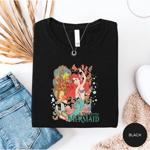 a black t-shirt with a colorful graphic of a mermaid character, along with a pair of blue jeans and a necklace.