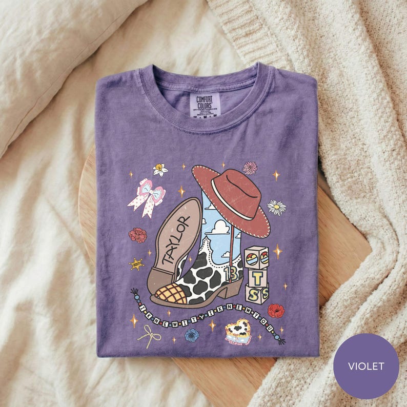 A purple t-shirt with a cartoon-style illustration of a cowboy hat, a shoe, and various other objects, along with the text "TAYLOR" and "IT'S" printed on it.