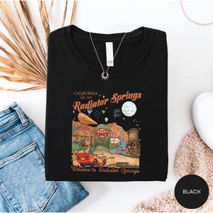 a black t-shirt with a graphic design depicting a desert landscape, a red car, and a moon, along with the text "California Est. 2012 Radiator Springs Welcome to Radiator Springs".