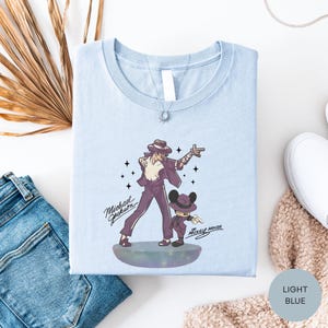 a light blue t-shirt with a graphic design of a man and a child, along with the text "Michael Jackson" and " Mickey Mouse". The t-shirt is laid out on a white surface, accompanied by a pair of blue jeans, a white scarf, and a white shoe.