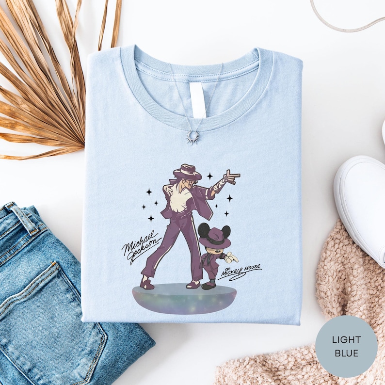 a light blue t-shirt with a graphic design of a man and a child, along with the text "Michael Jackson" and " Mickey Mouse". The t-shirt is laid out on a white surface, accompanied by a pair of blue jeans, a white scarf, and a white shoe.
