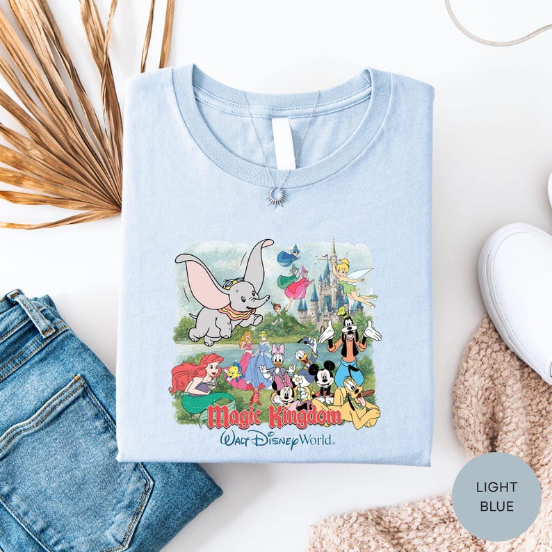 A light blue t-shirt with a Disney-themed graphic design, including characters from the Magic Kingdom, is displayed alongside a pair of jeans and a sweater.