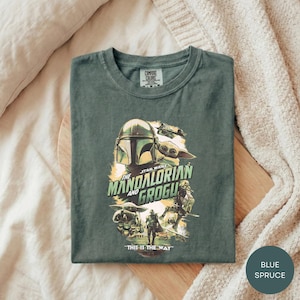 A green t-shirt with a graphic design featuring characters from the Star Wars series, specifically The Mandalorian and the Frog, laid out on a wooden surface.