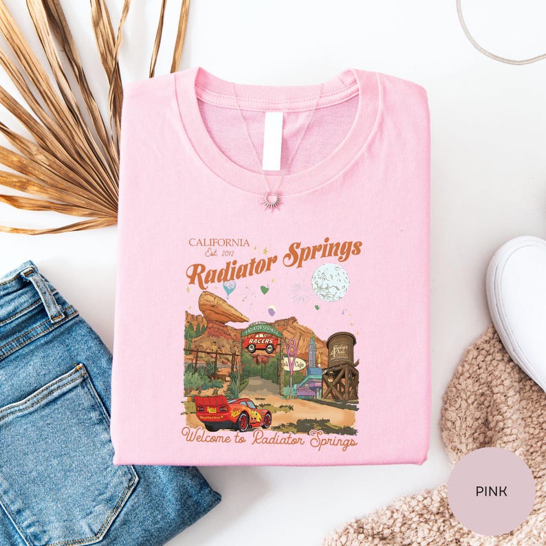 a pink t-shirt with a graphic design of a car and a desert landscape, along with text that says "California Radiator Springs Welcome to Radiator Springs".