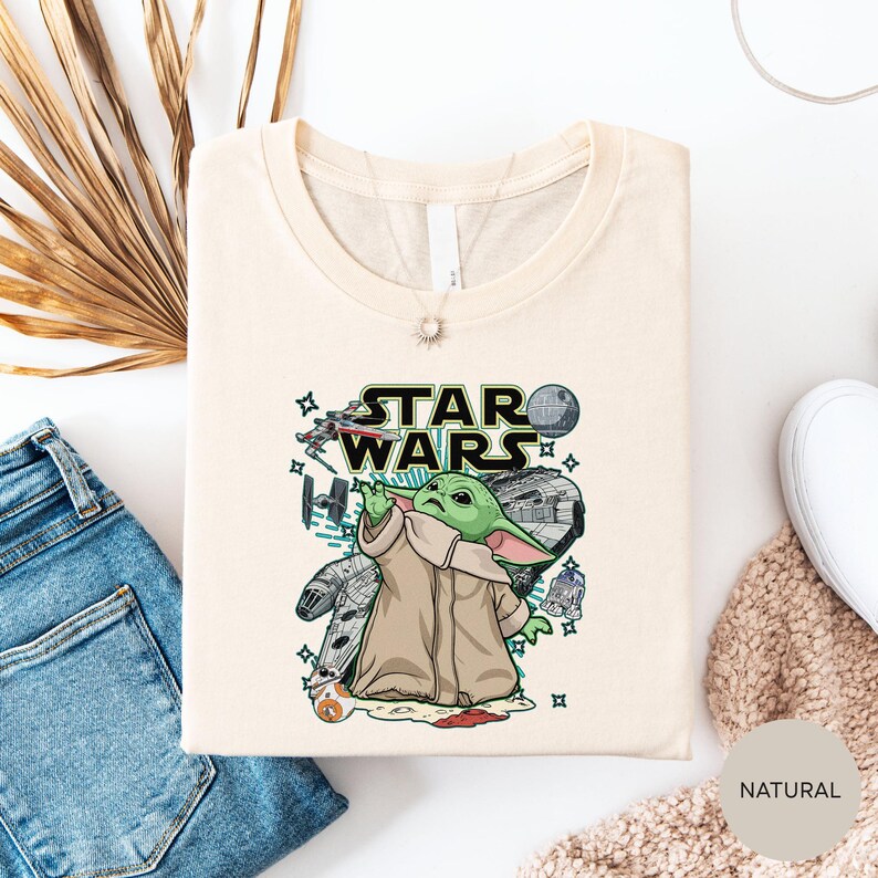 a white t-shirt with a Star Wars graphic design, including a depiction of the character Baby Yoda. The t-shirt is laid out on a white surface, along with a pair of jeans and a beige sweater.