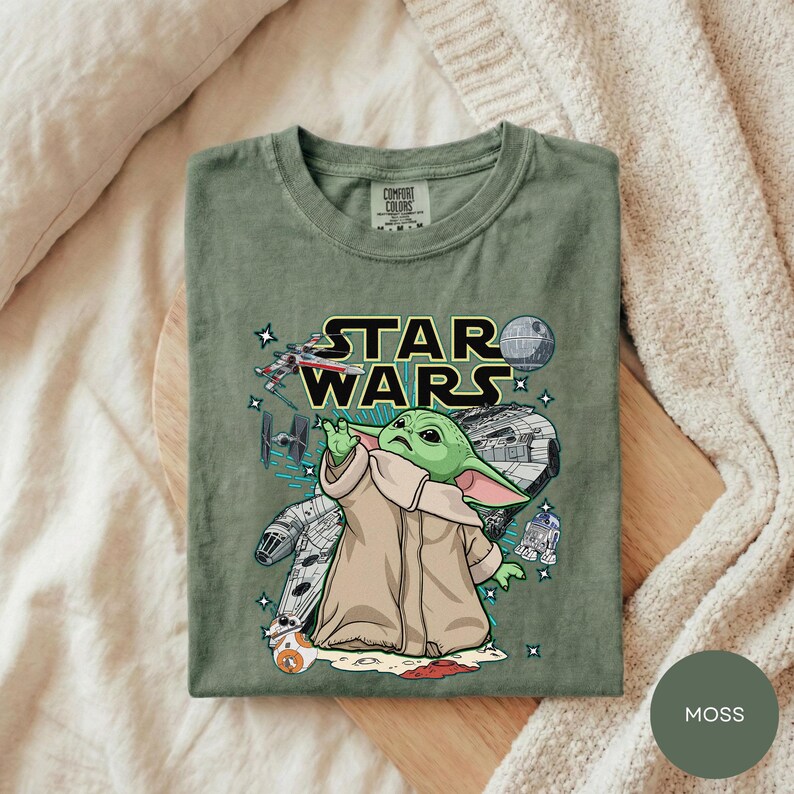 A green t-shirt with a Star Wars graphic design, featuring a young alien character, is laid out on a bed with a white blanket.