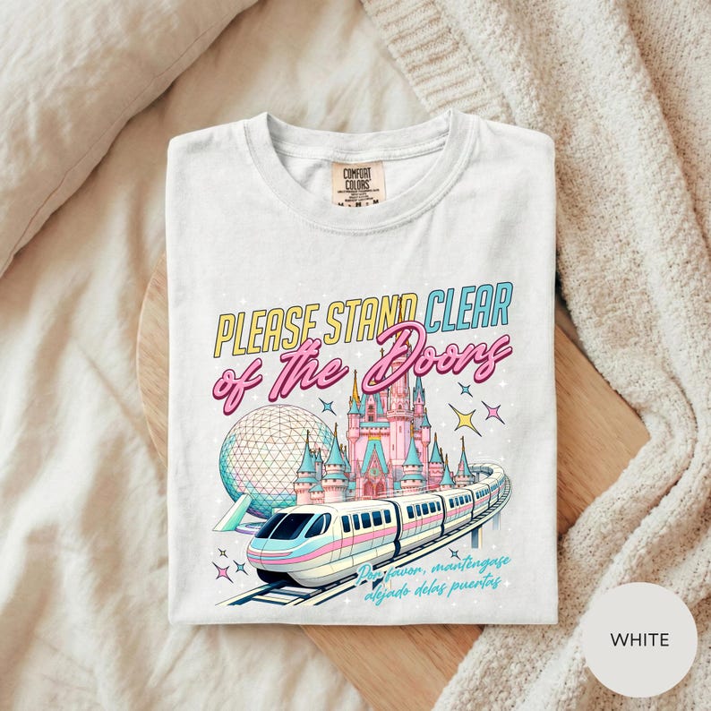 A white t-shirt with a colorful graphic design depicting a train and a castle, along with the text "Please Stand Clear of the Doors" and "Disneyland".