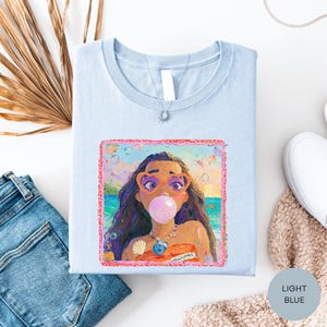 a light blue t-shirt with a colorful graphic of a young girl blowing a bubble gum bubble, surrounded by various clothing items and accessories.