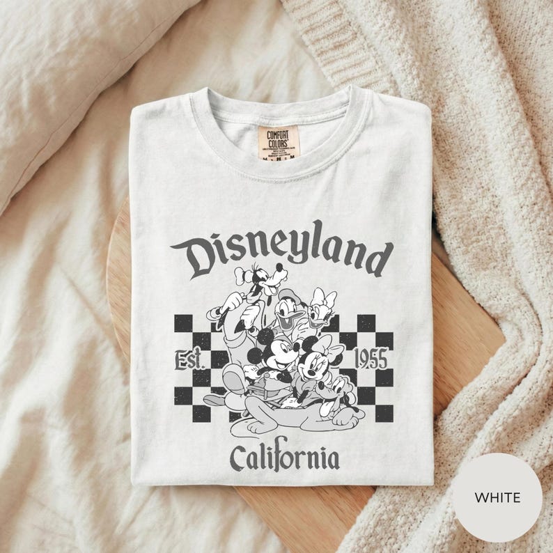 A white t-shirt with a graphic design of Disney characters and text, laid out on a wooden surface.