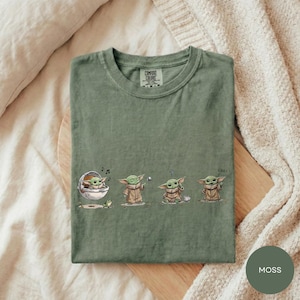 A green t-shirt with a graphic design depicting four baby alien characters from the Star Wars series, each in a different pose.