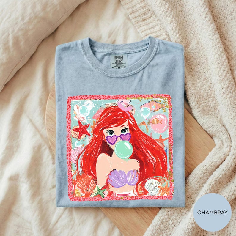 A blue t-shirt with a colorful graphic of a mermaid character blowing a bubble, surrounded by seashells and starfish.
