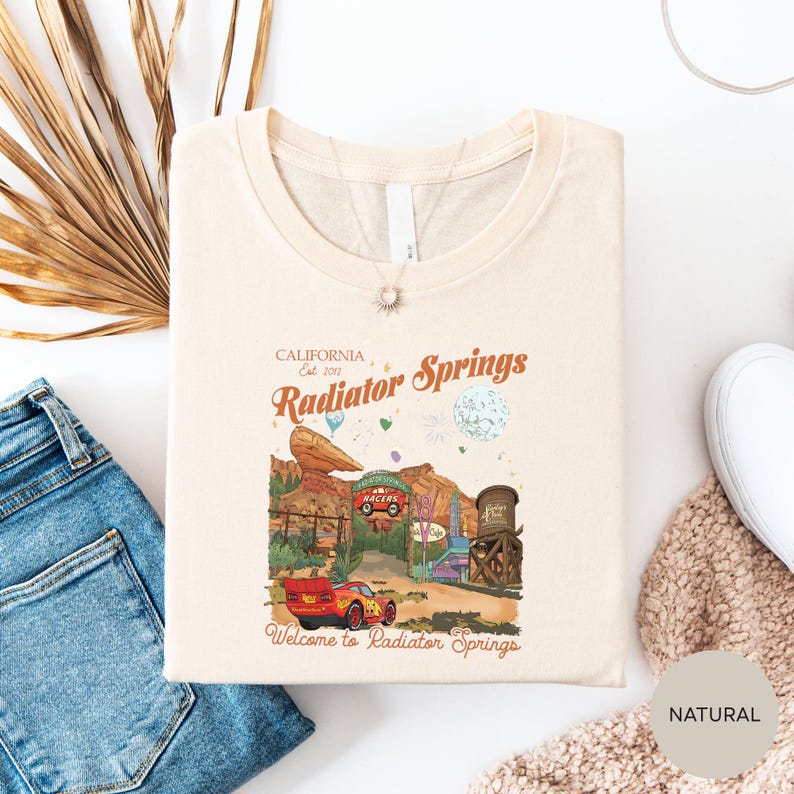 a white t-shirt with a graphic design depicting a desert landscape with a car and a building, along with the text "California Radiator Springs Welcome to Radiator Springs". The t-shirt is laid out on a white surface, with a pair of blue jeans and a beige sweater visible in the background.