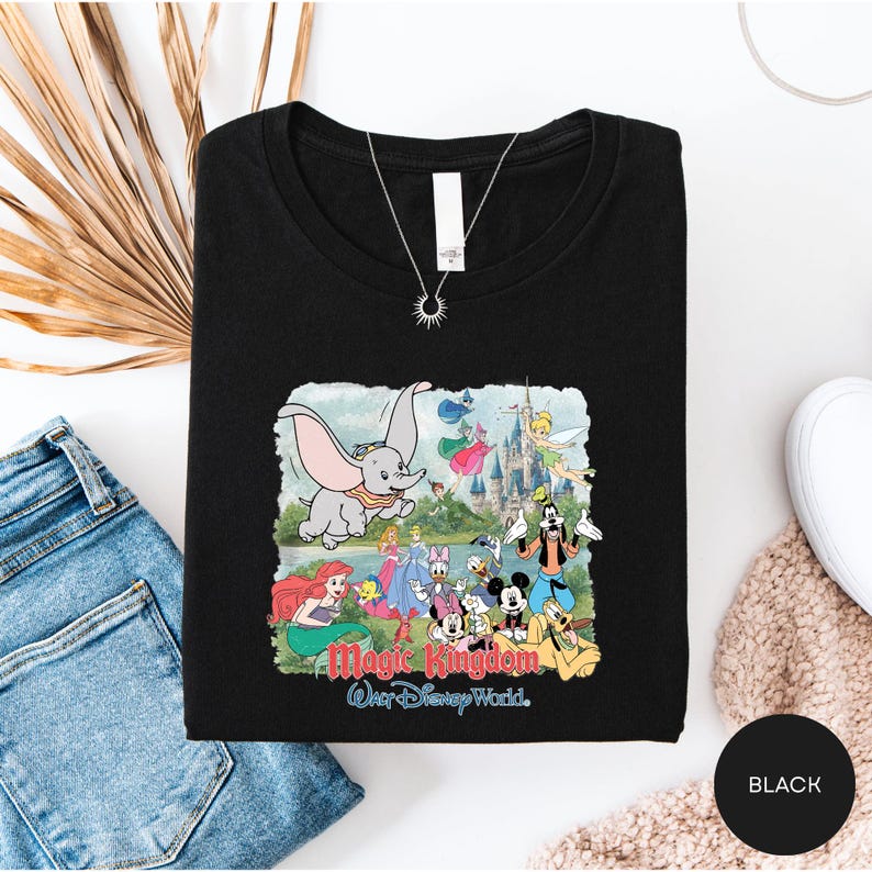 A black t-shirt with a Disney-themed graphic design, including characters from the Disney animated films, is displayed alongside a pair of jeans and a necklace.