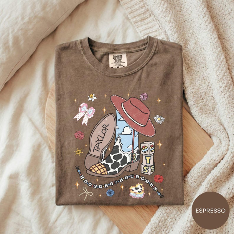A brown t-shirt with a cartoon-style design featuring a cowboy hat, a cow, and various other icons, along with the word "TAYLOR" printed on it.