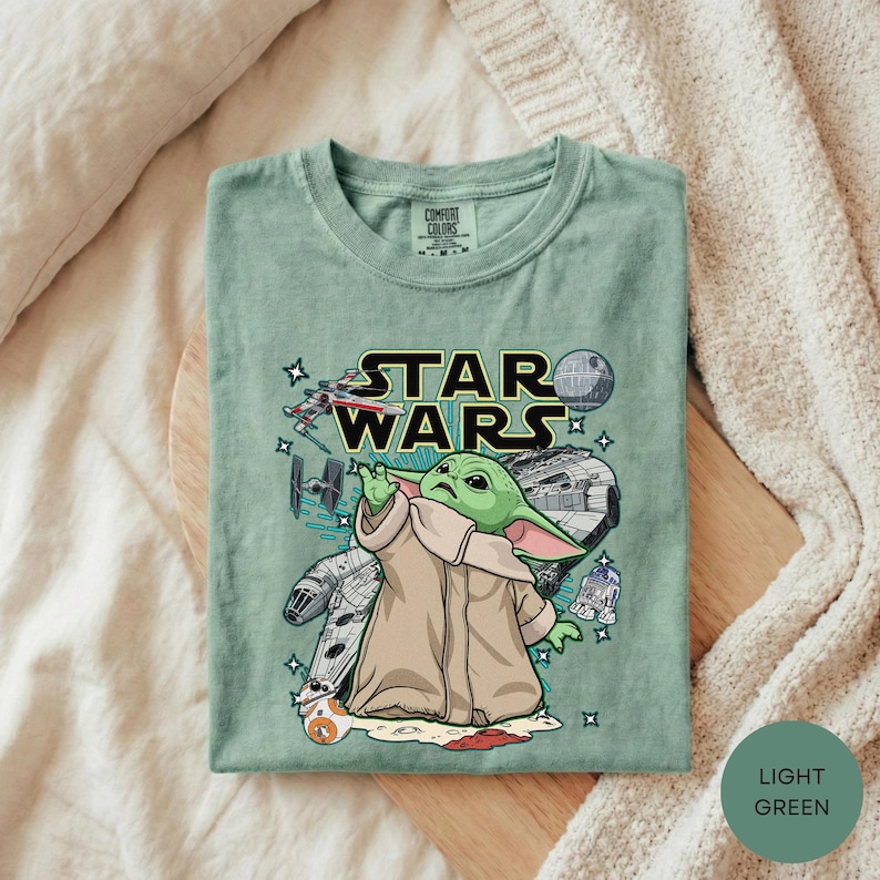 A green t-shirt with a Star Wars graphic design, featuring a young alien character, is laid out on a bed.