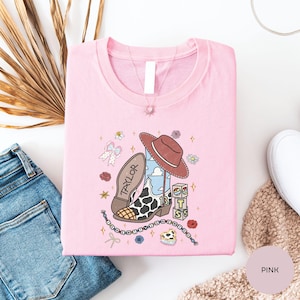 a pink t-shirt with a cartoon character and various accessories, such as a cowboy hat, a chain, and a flower, all set against a white background.