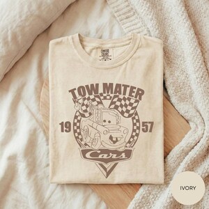 A beige t-shirt with a graphic design featuring a tow truck and the text "TOW MATTER Cars" printed on it, laid out on a wooden surface.