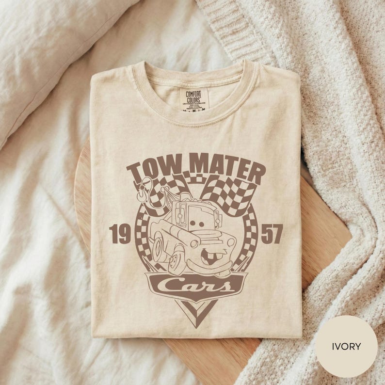 A beige t-shirt with a graphic design featuring a tow truck and the text "TOW MATTER Cars" printed on it, laid out on a wooden surface.