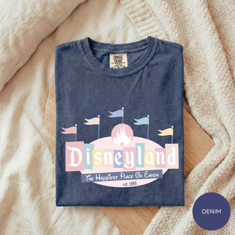 A blue t-shirt with a Disneyland logo and text, laid out on a bed with a blanket.