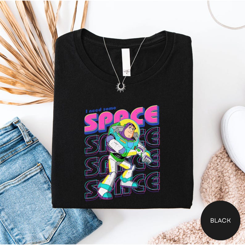 a black t-shirt with a colorful graphic of a character from the Toy Story franchise, along with the text "I need some space" printed on it. The t-shirt is laid out on a white surface, accompanied by a pair of blue jeans and a beige sweater.