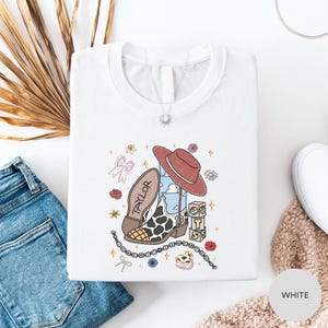 a white t-shirt with a colorful graphic design, including a cowboy hat, a cow, and various other cartoon-style elements. The t-shirt is laid out on a white surface, along with a pair of blue jeans and a beige sweater.