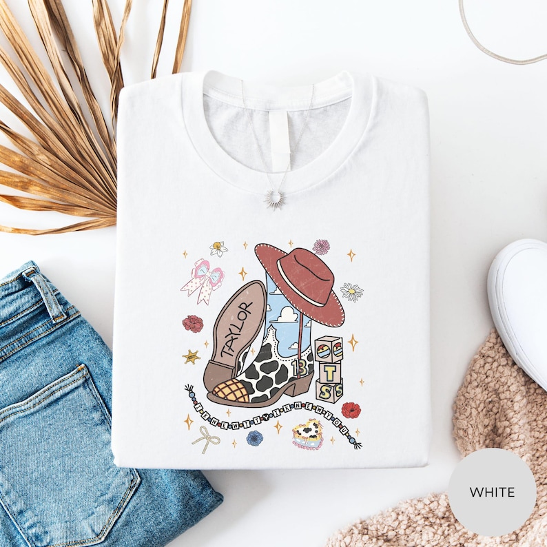 a white t-shirt with a colorful graphic design, including a cowboy hat, a cow, and various other cartoon-style elements. The t-shirt is laid out on a white surface, along with a pair of blue jeans and a beige sweater.
