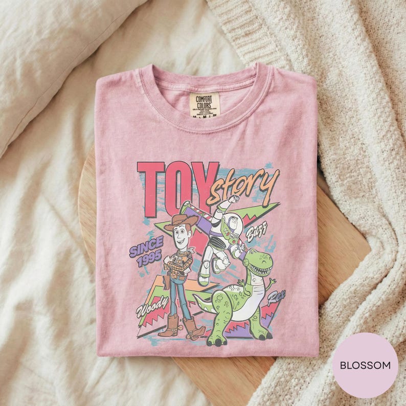 A pink t-shirt with a graphic design featuring characters from the Toy Story movie, including Woody and Buzz Lightyear, along with the text "TOY STORY SINCE 1995" and "Woody & Buzz".