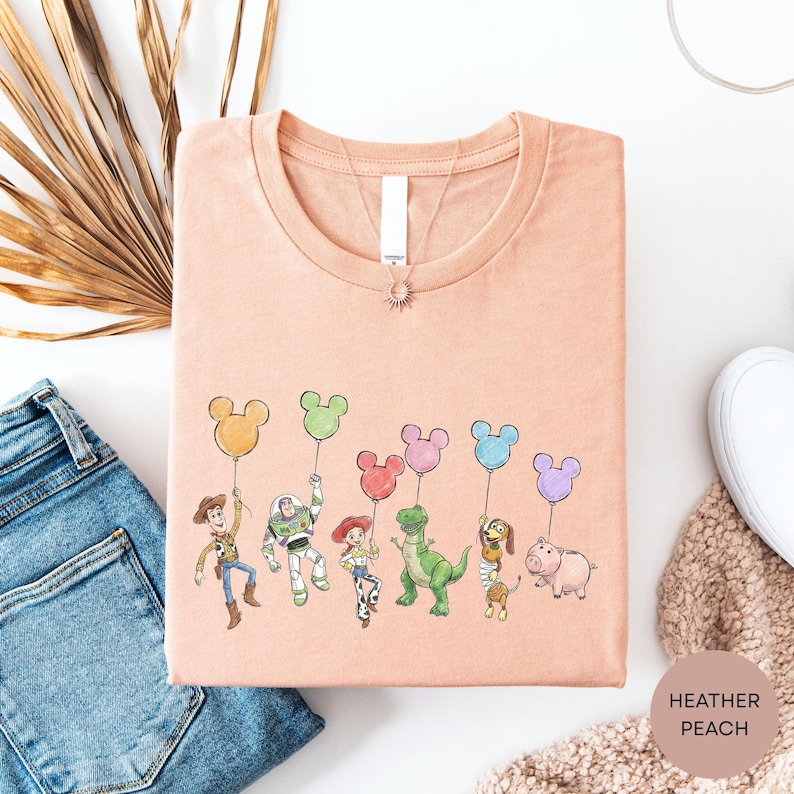 a pink t-shirt with a cartoon illustration of various Disney characters, including Woody, Buzz Lightyear, Timmy, and others, holding Mickey Mouse balloons. The t-shirt is laid out on a white surface, along with a pair of blue jeans and a beige sweater.