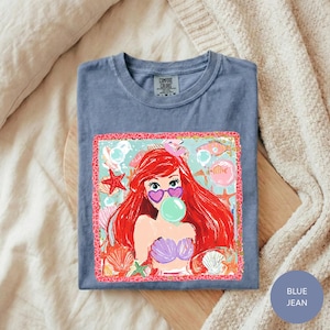 A blue t-shirt with a colorful graphic of a mermaid character blowing a bubble gum bubble, surrounded by seashells and starfish.