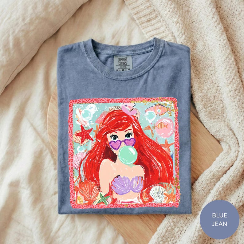 A blue t-shirt with a colorful graphic of a mermaid character blowing a bubble gum bubble, surrounded by seashells and starfish.