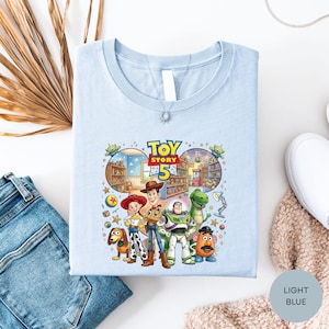 a light blue t-shirt with a colorful graphic of characters from the Toy Story movie, including Woody, Buzz Lightyear, and other beloved characters.