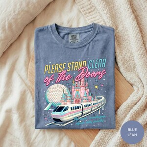 A blue t-shirt with a graphic design depicting a train and a castle, along with the text "Please Stand Clear of the Doors" and "Disneyland" printed on it.
