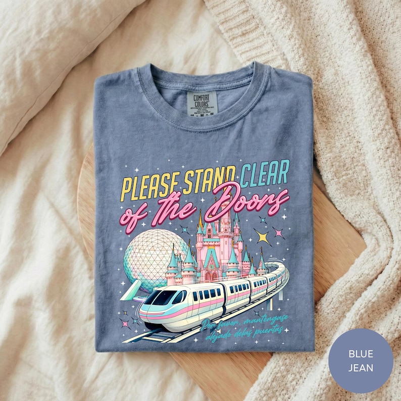 A blue t-shirt with a graphic design depicting a train and a castle, along with the text "Please Stand Clear of the Doors" and "Disneyland" printed on it.