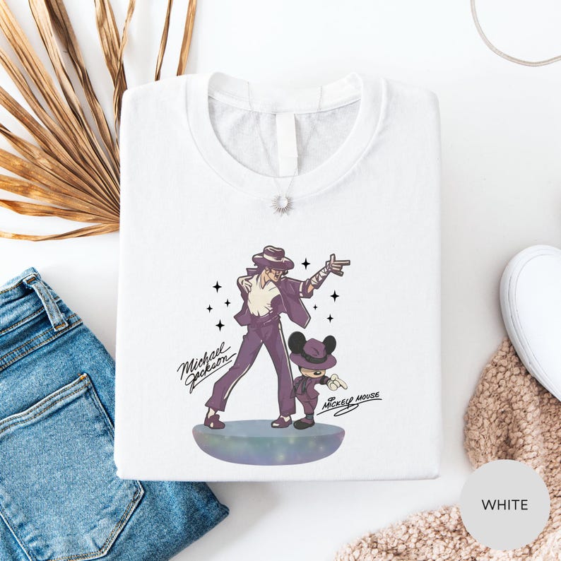 a white t-shirt with a graphic design of a man and a child, along with the text "Michael Jackson" and "Mickey Mouse". The t-shirt is laid out on a white surface, with a pair of blue jeans and a beige sweater visible in the background.