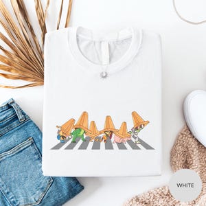 a white t-shirt with a graphic design of cartoon characters wearing orange hats, standing on a crosswalk.