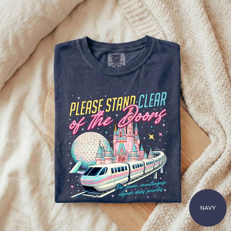 A navy blue t-shirt with a colorful graphic design depicting a train on tracks, a castle, and a globe, along with the text "Please Stand Clear of the Doors" and "Disneyland".