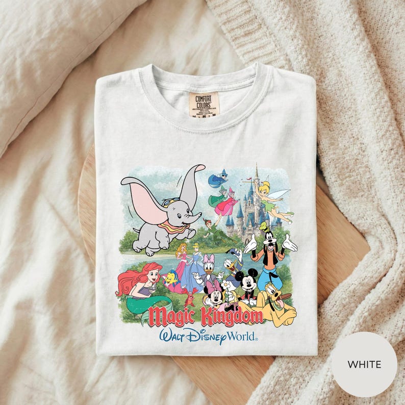 A white t-shirt with a colorful graphic design depicting various Disney characters from the Magic Kingdom, including Dumbo, Minnie Mouse, Mickey Mouse, Donald Duck, and other iconic figures.