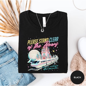 a black t-shirt with a colorful graphic design depicting a train and a castle, along with the text "Please stand clear of the doors" and "The doors are closing".