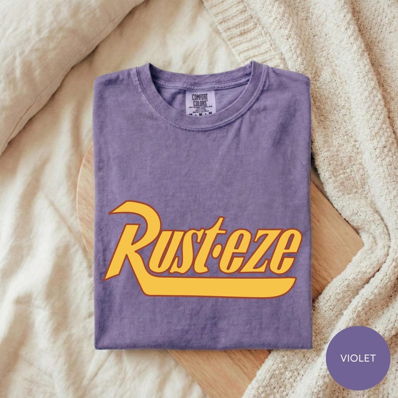 A purple t-shirt with the word "Rusteze" printed on it, laid out on a wooden surface.